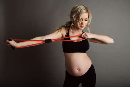 Pregnant beautiful blonde woman is wearing sportive black clothes doing exercises with rubber band isolated on gray, healthy life conceptの写真素材
