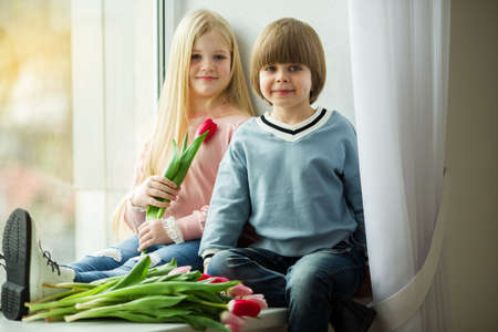 Pretty lovely kids are sitting on window sill , little boy and cute girl with bouquet of pink tulip flowers. Easter conceptの写真素材