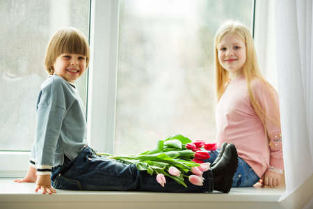 Pretty lovely kids are sitting on window sill , happy little boy and cute girl with bouquet of pink tulip flowers. Easter conceptの写真素材