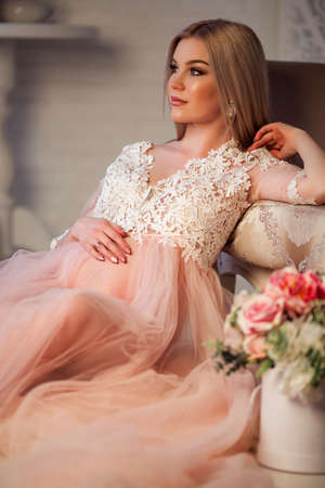 Beautiful pregnant young woman is wearing luxury pink lingerie dress at home in cozy interior with bouquet of flowersの写真素材