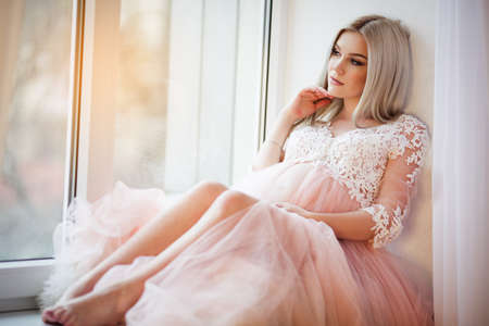 Beautiful pregnant young woman is wearing luxury pink lingerie dress at home sitting on window sill, spring timeの写真素材