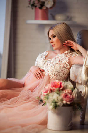 Pretty pregnant young woman is wearing luxury pink lingerie dress at home in cozy interior with bouquet of flowersの写真素材