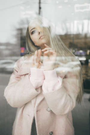 Unhappy pretty blonde girl is wearing pink coat behind the window, spring timeの写真素材
