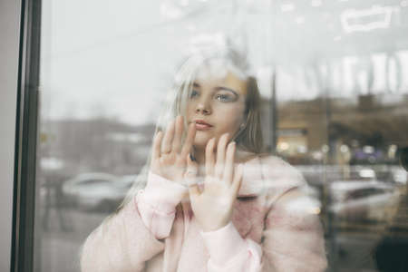 Sad pretty blonde girl is wearing pink coat behind the window, spring timeの写真素材