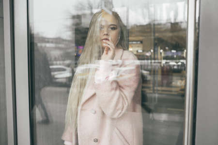 Sad lonely pretty blonde girl is wearing pink coat behind the window, spring time