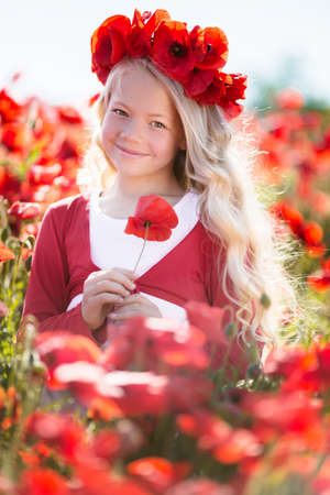 Beautiful blonde child girl is wearing wreath from red flowers in poppy meadow, spring timeの写真素材