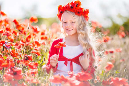 Beautiful blonde child girl is wearing wreath from red flowers in poppy meadow, spring timeの写真素材