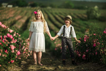 Cute child boy and girl are holding hands are standing in pink rose field. Wearing stylish clothes, summer timeの写真素材