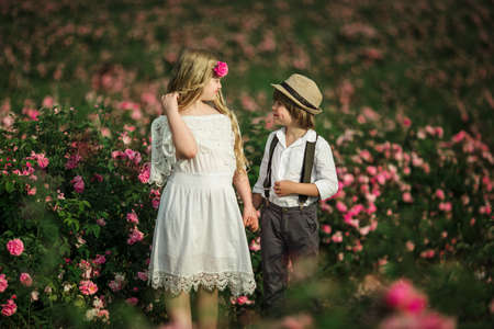 Cute child boy and girl are holding hands are standing in pink rose field. Wearing stylish clothes, summer timeの写真素材