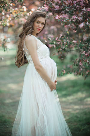 Pregnant pretty woman is wearing white fashion dress posing in pink apple garden, summer timeの写真素材