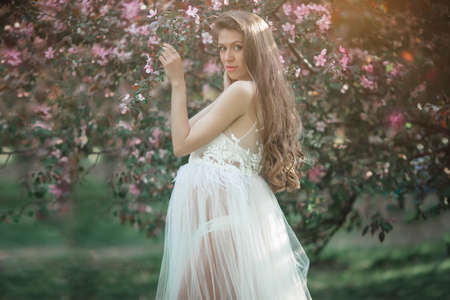 Pregnant pretty woman is wearing white fashion dress posing in pink blossom apple garden, summer timeの写真素材