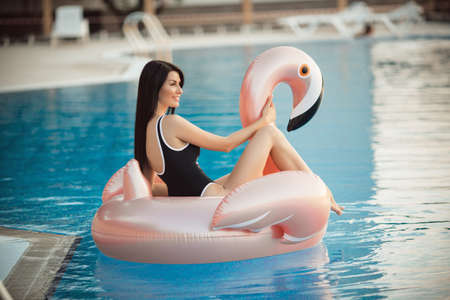 Stunning sexy woman is wearing black bikini sitting in swimming pool with blue water on a pink...