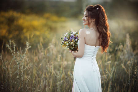 Beautiful young bride with bouquet of wildflowers in field over sunset lightsの写真素材
