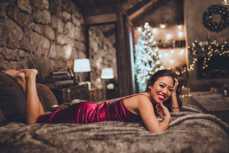 Beautiful happy asian woman is sitting in bed at home near christmas tree in cozy interior. Interior with christmas decorations.の写真素材