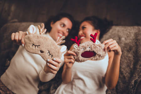 Two happy girls friends are lying in bed and wearing sleeping mask at home near christmas tree in cozy interior. Interior with christmas decorations.の写真素材