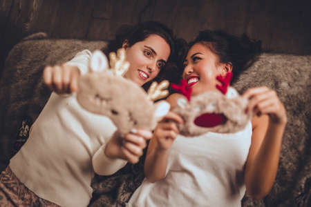 Two happy girls friends are lying in bed and wearing sleeping mask at home near christmas tree in cozy interior. Interior with christmas decorations.の写真素材