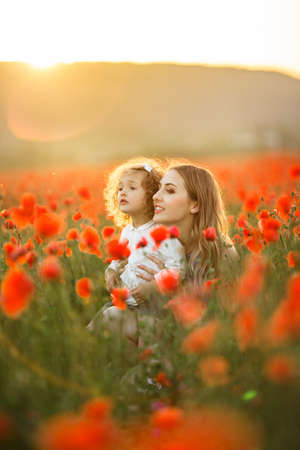 Beautiful smiling baby girl with mother are having fun in field of red poppy flowers over sunset lights, spring timeの写真素材