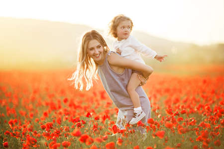 Beautiful smiling child girl with mother are having fun in field of red poppy flowers over sunset lightsの写真素材