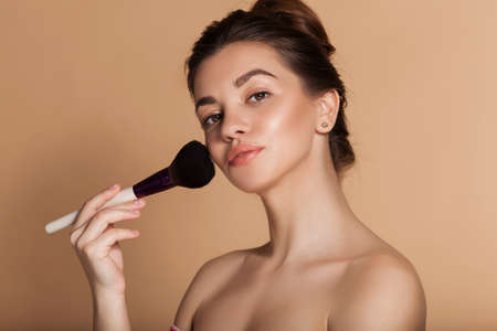 Closeup portrait of pretty girl with makeup brush in hand. Isolated on beige. Beauty and cosmetics concept.の写真素材