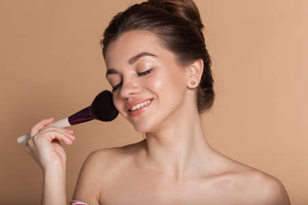Closeup portrait of beautiful young girl with perfect skin is holding makeup brush in hand. Isolated on beige. Beauty and cosmetics concept.の写真素材