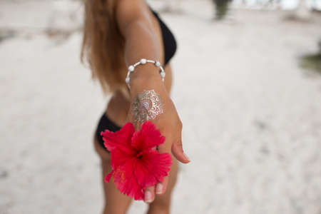 Young tanned woman with perfect body with golden tattoo on her hand and red flower is walking on the beach. Beauty and fashion concept.の写真素材