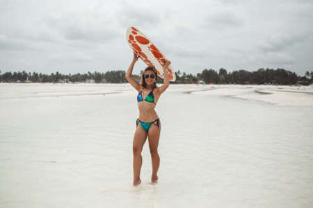 Young sexy woman with sportive tanned body, smiling and jogging with surf board on the beach in cloudly summer day during low tide in Zanzibarの写真素材