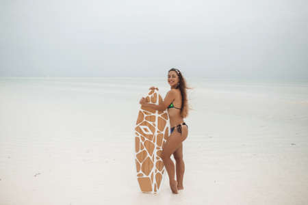 Young sexy woman with sportive tanned body, smiling and jogging with surf board on the beach in cloudly summer day during low tide in Zanzibarの写真素材