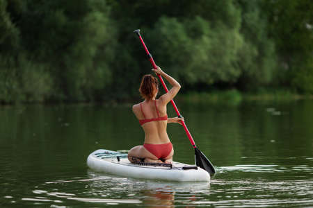 Young pretty fitness girl in swimwear is paddling on a SUP in the middle of the lake with a forest in the background.の写真素材