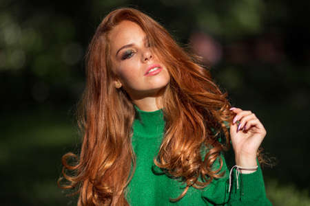Sunshine young smiling woman with red curly hair is wearing green sweater in autumn park.の写真素材