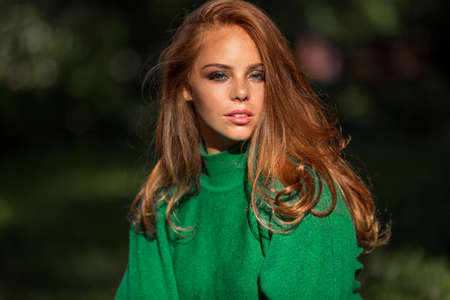Sunshine young smiling woman with red curly hair is wearing green sweater in autumn park.の写真素材