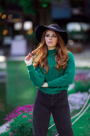 Sunshine young teen girl with red curly hair is wearing green knitted sweater and fashion hat in autumn park with flowers.の写真素材
