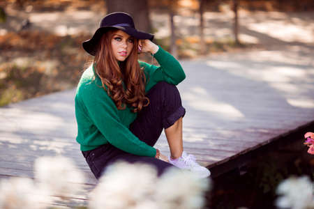 Portrait of sunshine young teen girl with red curly hair is wearing green sweater and fashion hat is sitting on bridge in autumn park.の写真素材