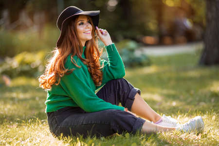 Sunshine young teen girl with red curly hair is wearing green knitted sweater and hat is sitting on grass in autumn park.の写真素材