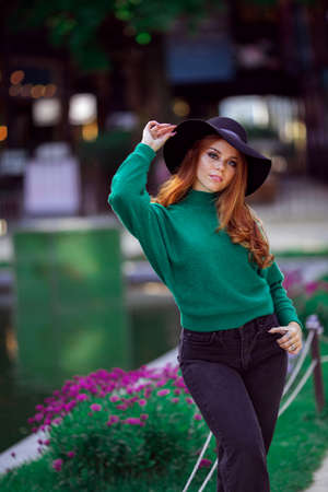 Sunshine young teen girl with red curly hair is wearing green knitted sweater and fashion hat in autumn park.の写真素材