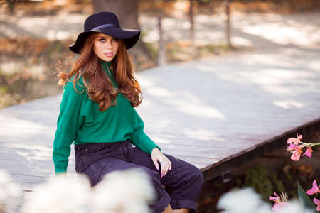 Portrait of sunshine young teen girl with red curly hair is wearing green sweater and fashion hat is sitting on bridge in autumn park.の写真素材