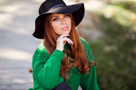 Portrait of sunshine young teen girl with red curly hair is wearing green sweater and fashion hat in autumn park.の写真素材