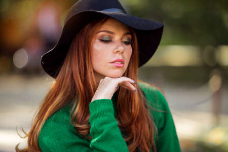 Sunshine young teen girl with red curly hair is wearing green sweater and hat in autumn park.の写真素材