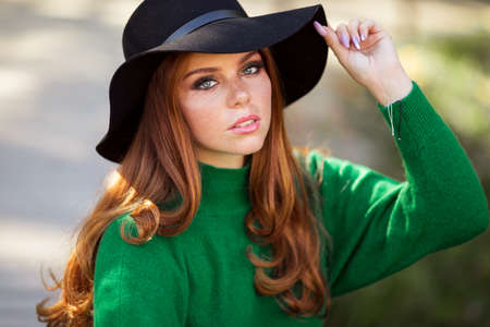 Portrait of sunshine young teen girl with red curly hair is wearing green sweater and fashion hat in autumn park.の写真素材