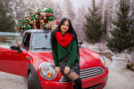 Pretty young girl is sitting on red car with decorated xmas tree on the roof, holiday concept.の写真素材