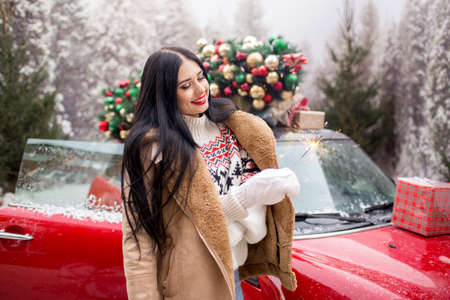 Beautiful young girl is wearing winter clothes holding sparklers near red car with decorated xmas tree on the roof, holiday and new years concept.の写真素材