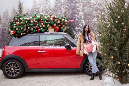 Beautiful young girl is holding gift box near red car with decorated xmas tree on the roof, holiday and new years concept.の写真素材