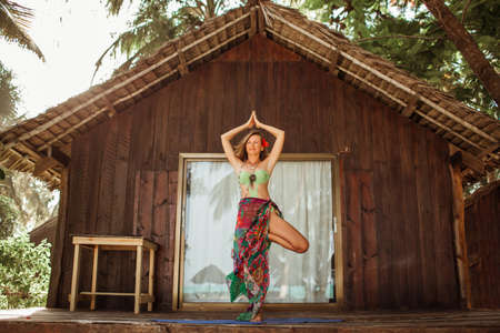 Yoga asana by young beautiful woman near wooden house in the tropical paradise with palms and banana trees on Zanzibar islandの写真素材