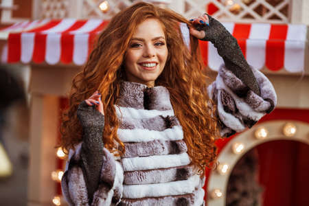 Outdoor portrait of young beautiful redhead happy smiling girl is wearing fur coat near Christmas decorations on Red Square in Moscow.の写真素材