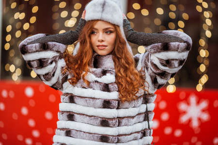 Outdoor portrait of young beautiful redhead happy smiling girl is wearing fur warm hat. Festive...