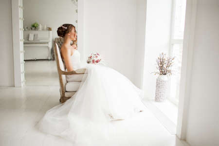 Young bride is wearing beautiful dress holding a bouquet of flowers in bright white studio. Wedding concept.の写真素材