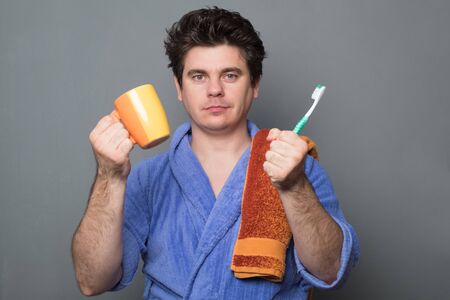 Sleepy man in a bathrobe brushes his teeth in the morning. Man in a bathrobe is holding a towel.