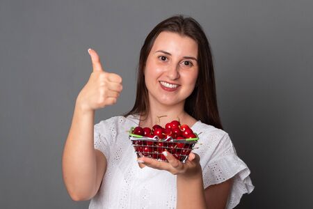 Positive woman holds a basket of cherries. Smiling girl in a summer dress holds a cherry.