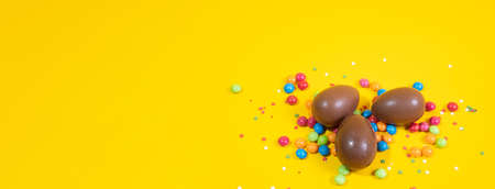 Banner. Chocolate Easter eggs with colored candies on a yellow background.の写真素材