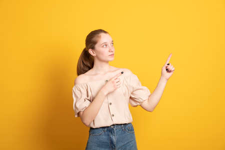 Smiling blond haired girl gives advice and points right, promots product, wears casual t-shirt, poses against yellow background, indicates at advertisement, introduces promo with pleased expressionの写真素材