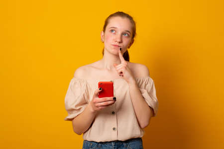Cute girl ponders on how to answer question, thinks deeply about something, uses modern mobile phone, tries to made up good message, keeps index finger near lips.の写真素材